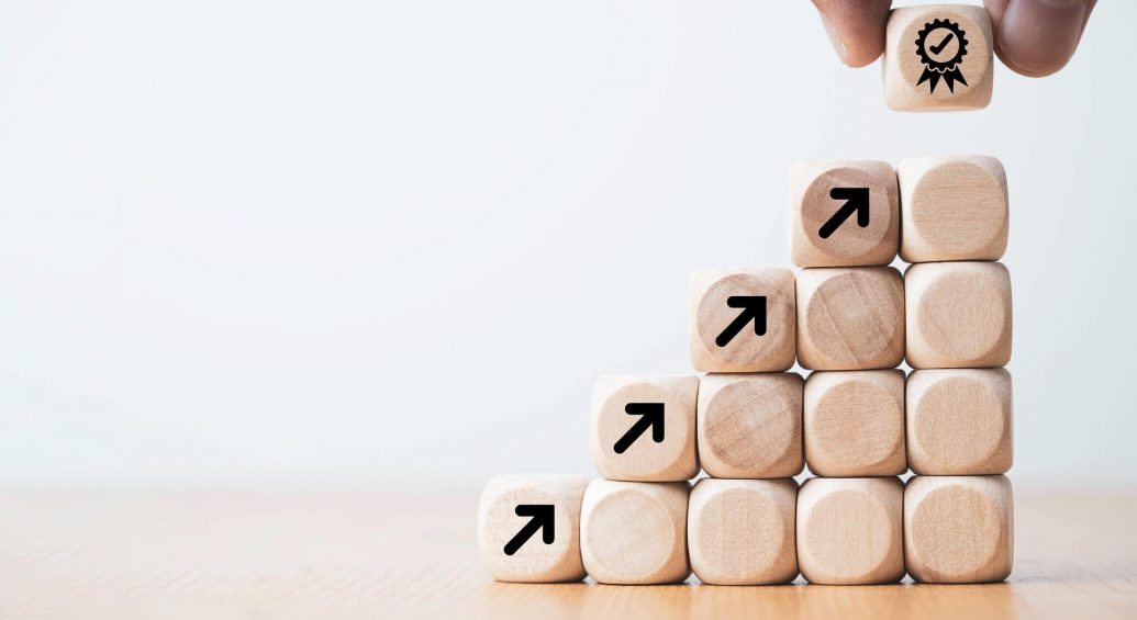 Hand placing a wooden cube with a ribbon icon on it on top of a set of staggered wooden cubes building upwards